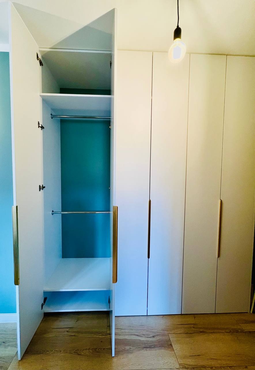 Biała szafa wnękowa z otwartymi drzwiami, widoczna półka i drążki na ubrania, wnętrze szafy w kolorze turkusowym, podłoga drewniana, oświetlenie żarówką edisona.