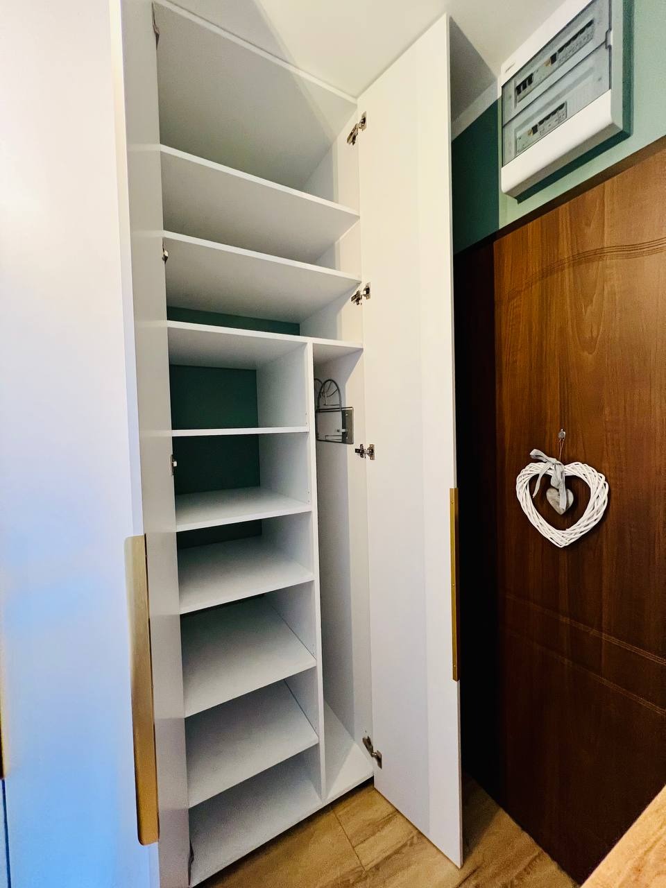 Otwarta, biała szafa wnękowa z półkami, widoczne złote uchwyty, na ścianie obok brązowe drzwi i dekoracja w kształcie serca.