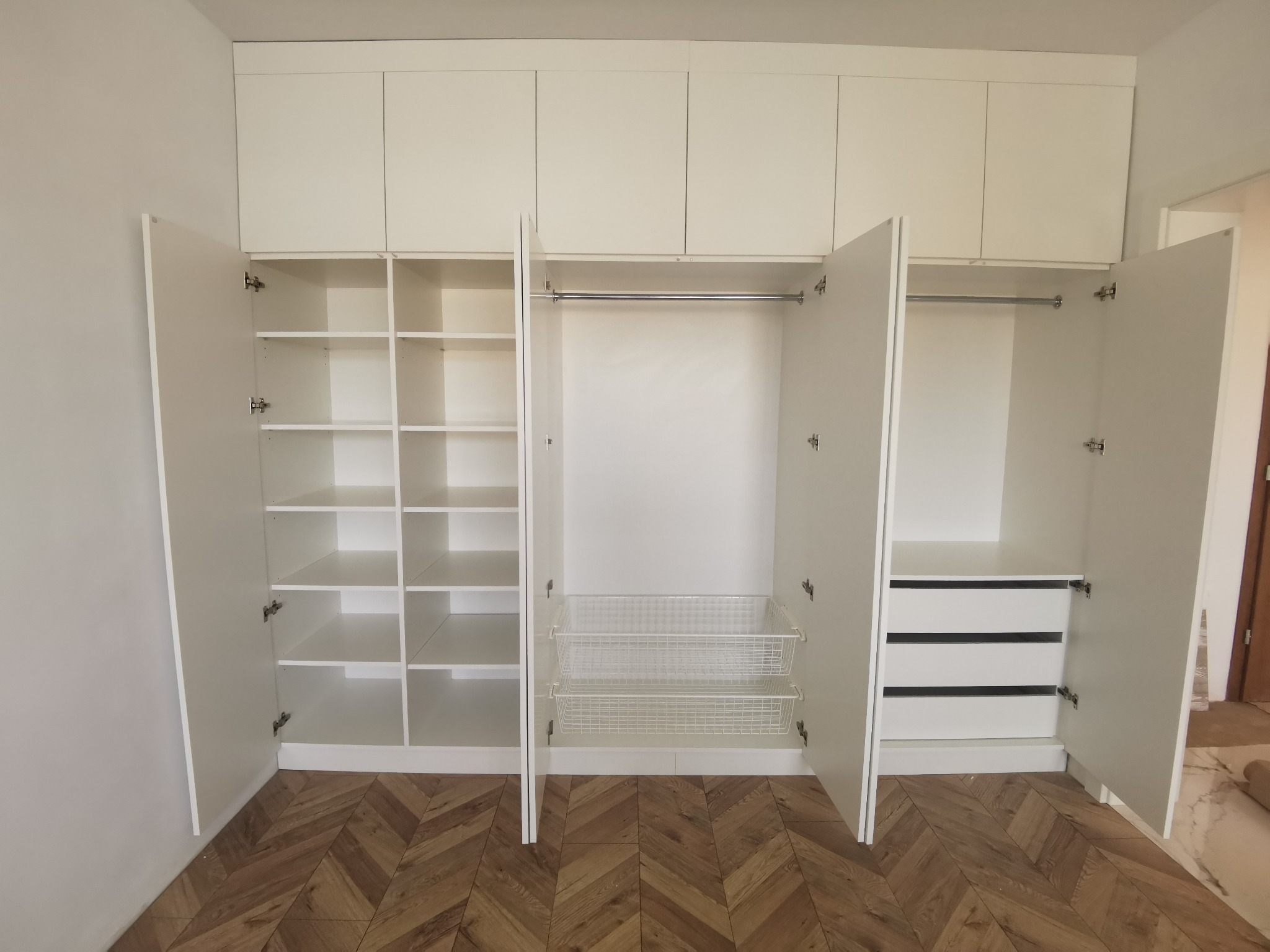 Biała szafa wnękowa z otwartymi drzwiami, ukazująca półki, kosze druciane i szuflady, na tle parkietu w jodełkę.