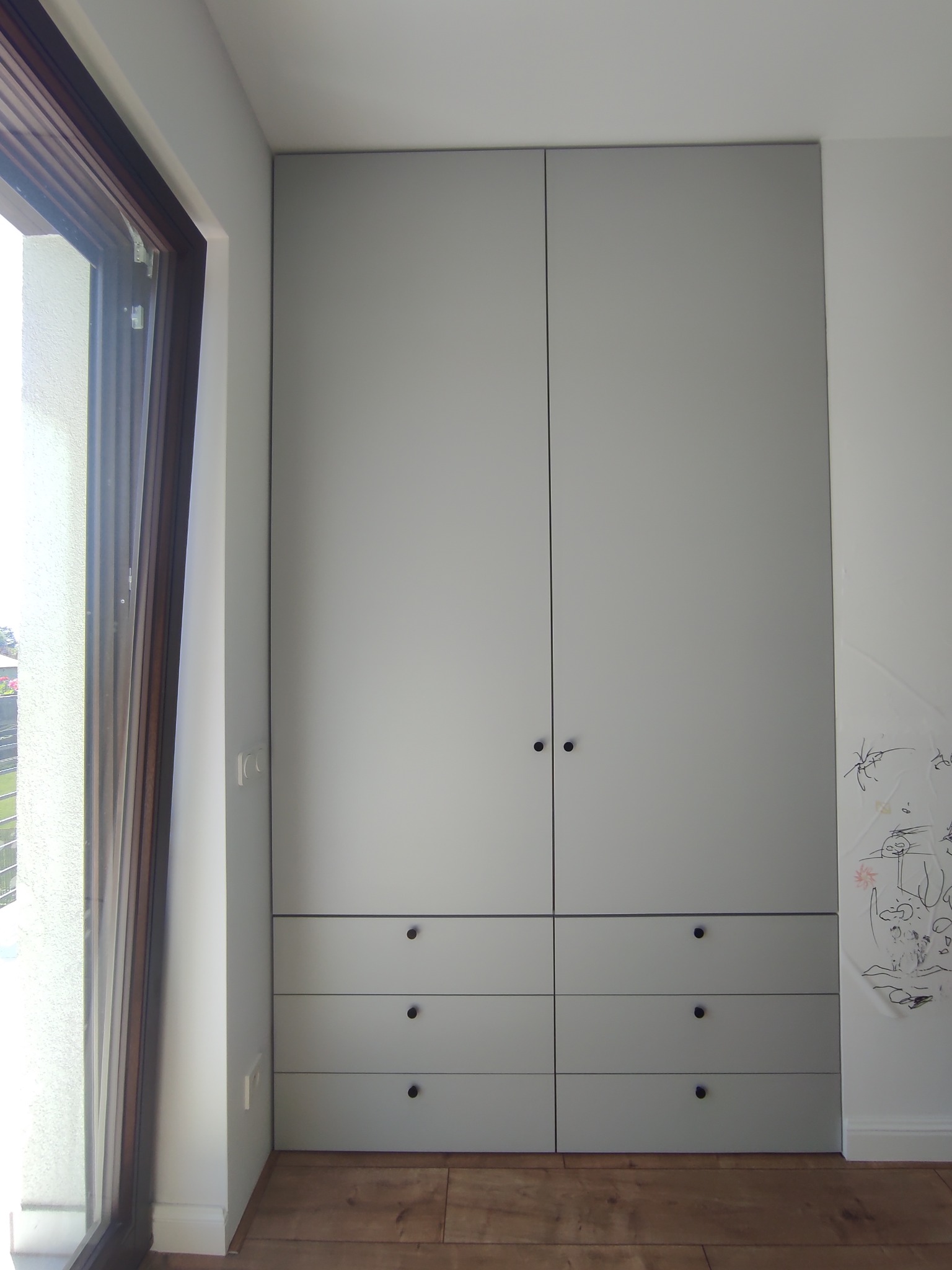 Szara szafa wnękowa z ciemnymi okrągłymi uchwytami, sześć szuflad u dołu, obok okna z widokiem na zewnątrz, na ścianie dziecięce rysunki.
