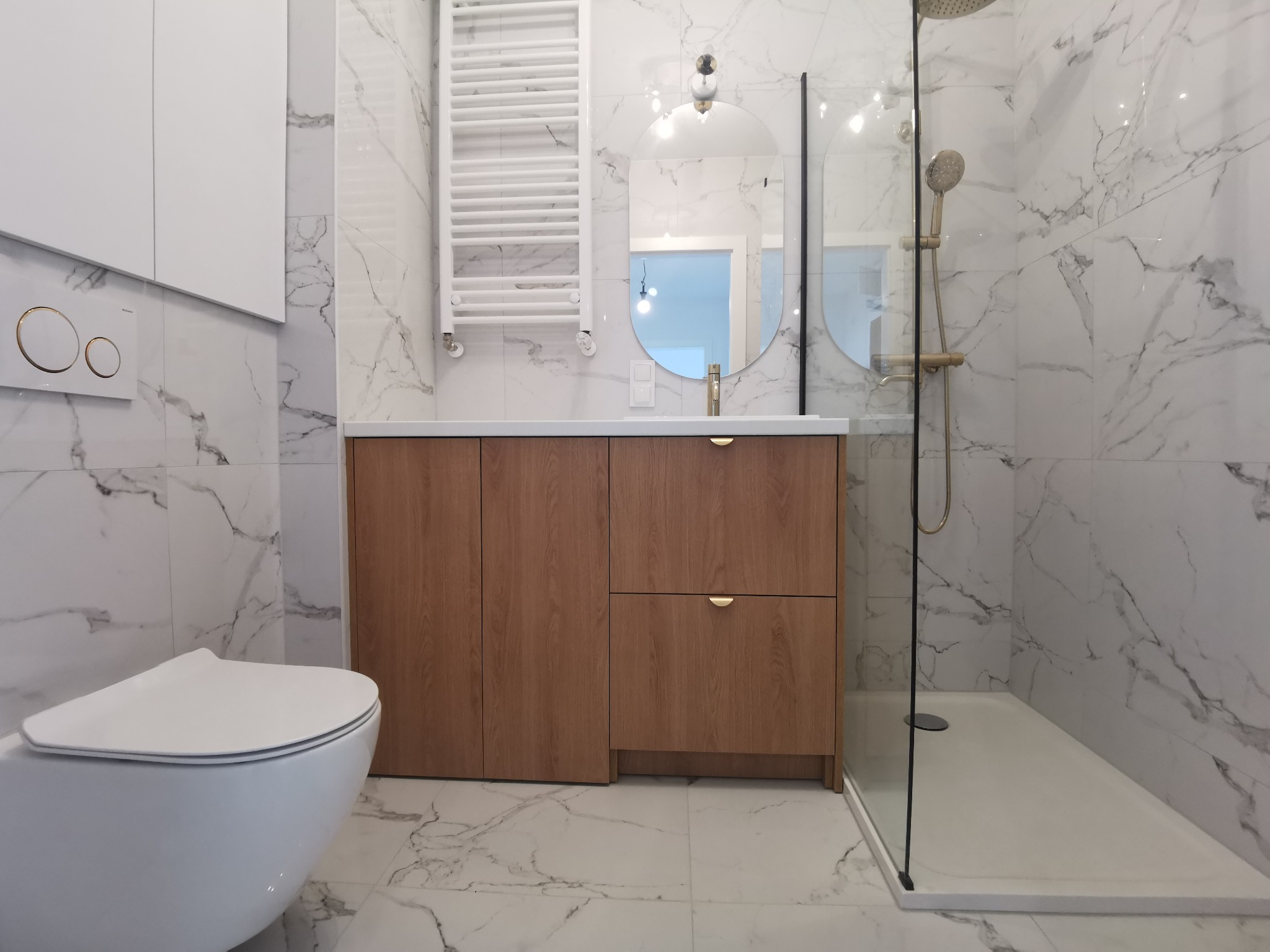 Nowoczesna łazienka z białymi marmurowymi płytkami, drewnianą szafką pod umywalką z okrągłymi złotymi uchwytami, owalnym lustrem, białą toaletą i kabiną prysznicową ze złotą armaturą.