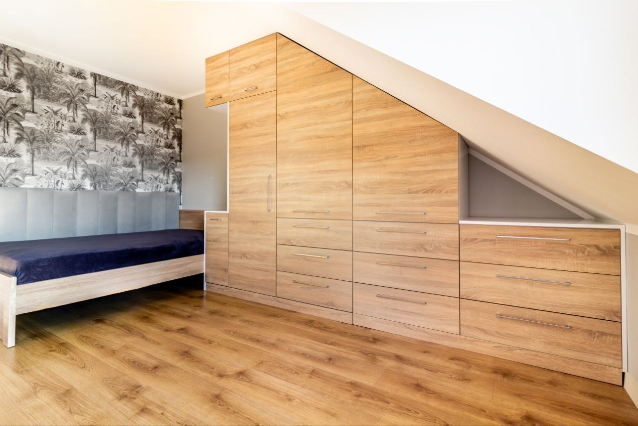 Drewniana szafa wnękowa z szufladami i szafkami, dopasowana do skośnego sufitu na poddaszu, z łóżkiem i tapetą w tropikalne wzory w tle.