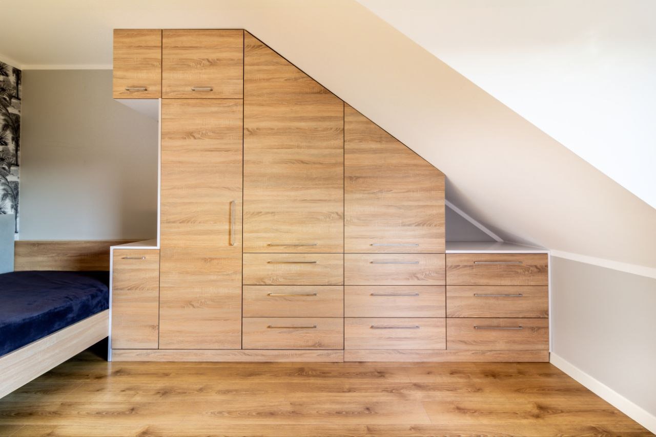 Drewniana szafa wnękowa na poddaszu we Wrocławiu, dopasowana do skosu dachu, zintegrowana z ramą łóżka, widoczna na tle ściany z tapetą.