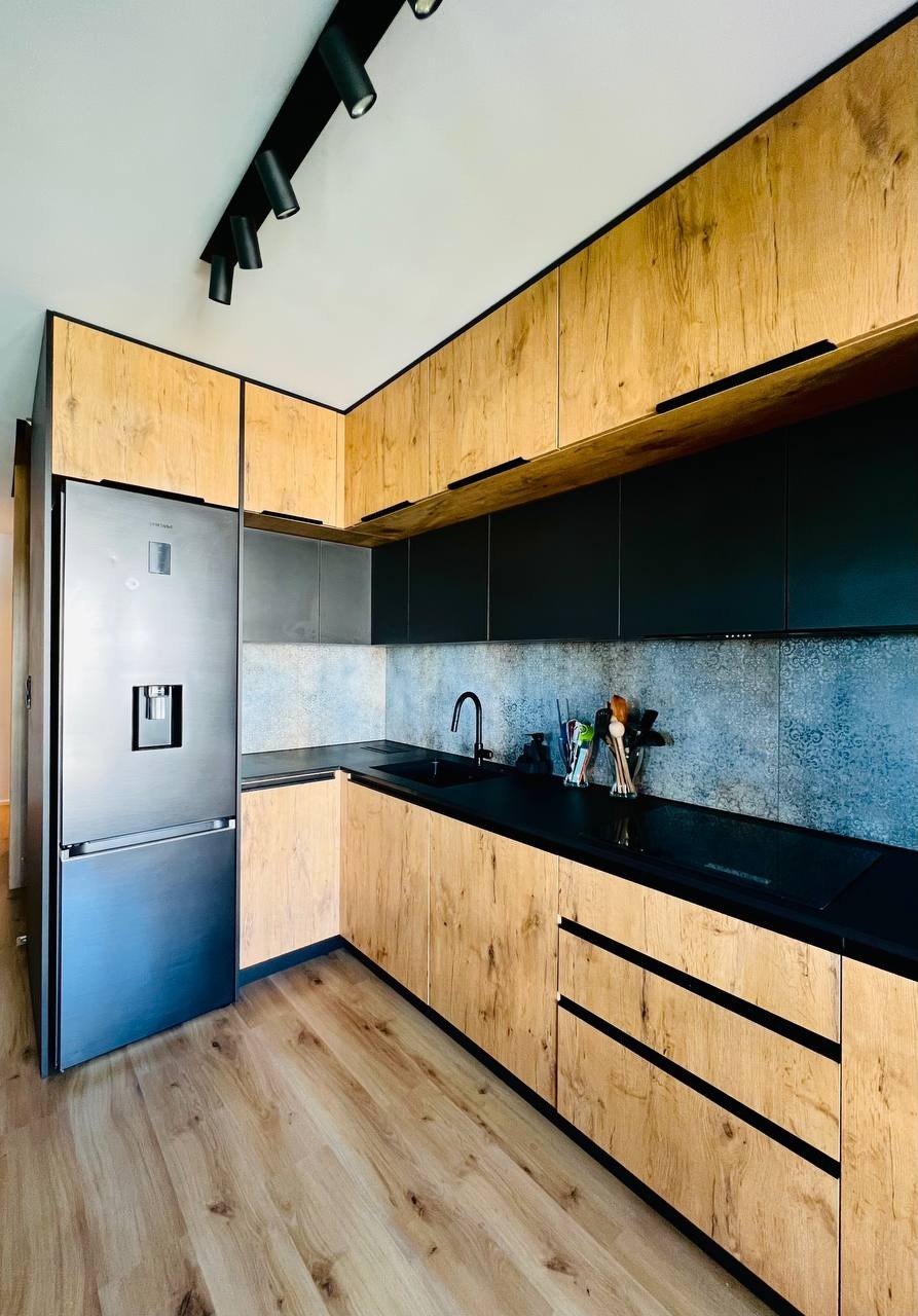 Nowoczesna kuchnia z drewnianymi frontami i czarnymi blatami, oświetlona punktowo, z widoczną lodówką z kostkarką i drewnianą podłogą.