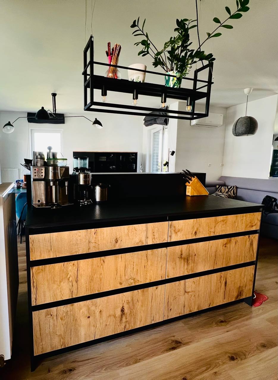 Wyspa kuchenna z drewnianymi frontami i czarnym blatem, na którym stoją ekspresy do kawy i przybornik na noże. Nad nią designerska lampa z roślinami.