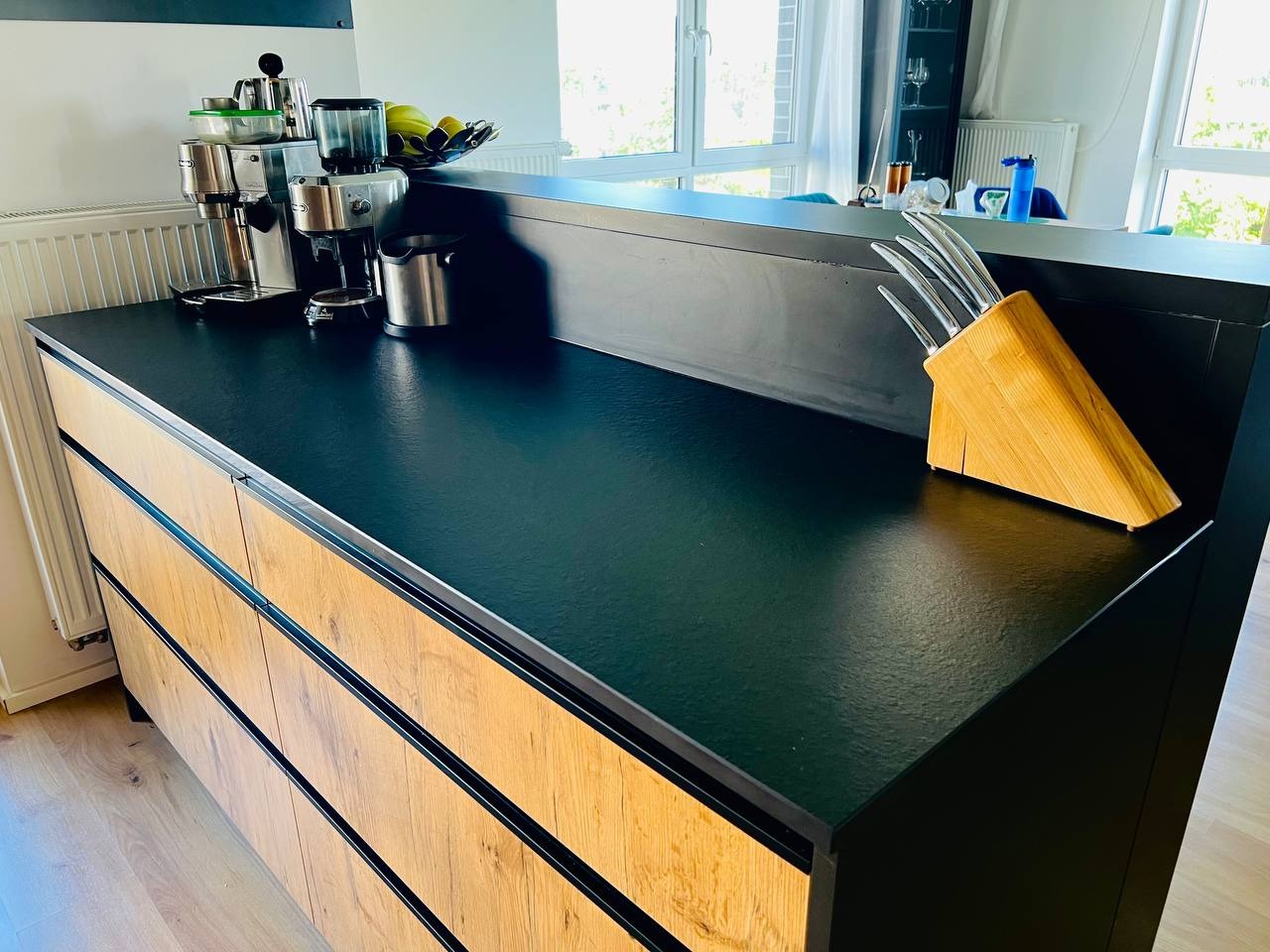 Nowoczesna, czarno-drewniana wyspa kuchenna z ekspresami do kawy i blokiem na noże, oświetlona naturalnym światłem w minimalistycznym wnętrzu.