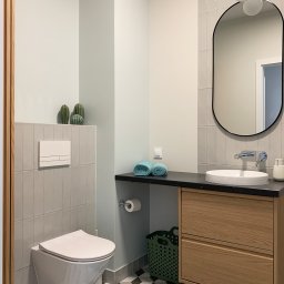 Hello Home Maja Maik - Jasna łazienka z geometryczną podłogą: toaleta podwieszana, szafka z umywalką, owalne lustro i dekoracyjne kaktusy. Minimalistyczny design z akcentami turkusu.