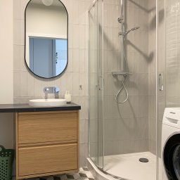 Hello Home Maja Maik - Jasna łazienka z kabiną prysznicową, umywalką na szafce z drewna i wzorzystą podłogą. Uwagę przykuwa owalne lustro z podświetleniem.