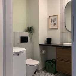 Hello Home Maja Maik - Nowoczesna łazienka w odcieniach szarości i zieleni z geometryczną podłogą, wiszącą toaletą i drewnianą szafką pod umywalką. Uwagę przykuwa roślina w czarnej doniczce.