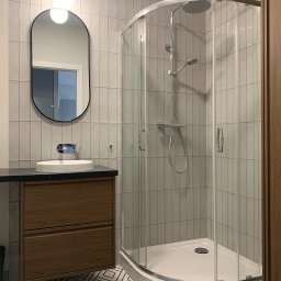 Hello Home Maja Maik - Nowoczesna łazienka z kabiną prysznicową narożną, umywalką na szafce z ciemnym blatem i owalnym lustrem podświetlanym. Geometryczny wzór na podłodze.