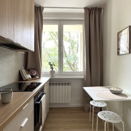 Hello Home Maja Maik - Jasna, wąska kuchnia z drewnianymi akcentami: zabudowa, blat, podłoga. Biały stół z krzesłami, okno z widokiem na zieleń. Minimalistyczny wystrój.