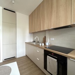 Hello Home Maja Maik - Jasna kuchnia w stylu skandynawskim: drewniane szafki górne, beżowe dolne, czarna płyta indukcyjna i piekarnik. Minimalistyczne oświetlenie sufitowe.