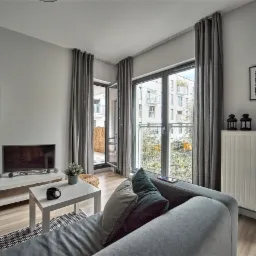 Jasne wnętrze salonu w neutralnych barwach z szarymi zasłonami, balkonem i widokiem na nowoczesne budynki, przygotowane do wynajmu długoterminowego w warszawskim Wilanowie.