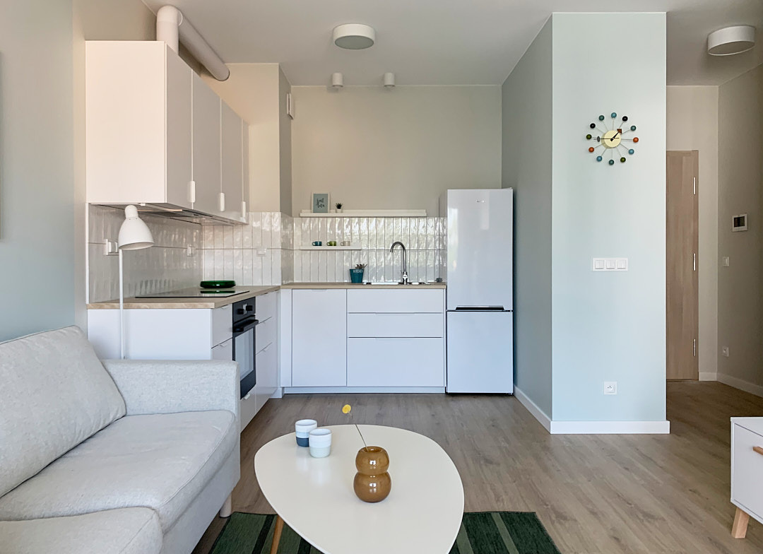 Jasne wnętrze z aneksem kuchennym: białe meble, drewniana podłoga, pastelowe ściany. Na ścianie zegar w stylu retro. Minimalistyczny design z akcentami kolorystycznymi.