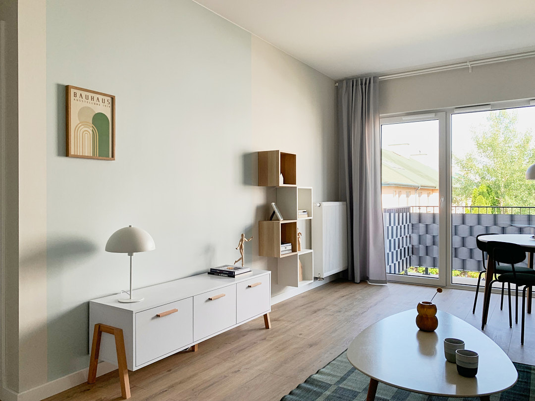 Jasne wnętrze mieszkania z drewnianą podłogą, białymi meblami i plakatem Bauhaus. Widok na balkon przez przeszklone drzwi.