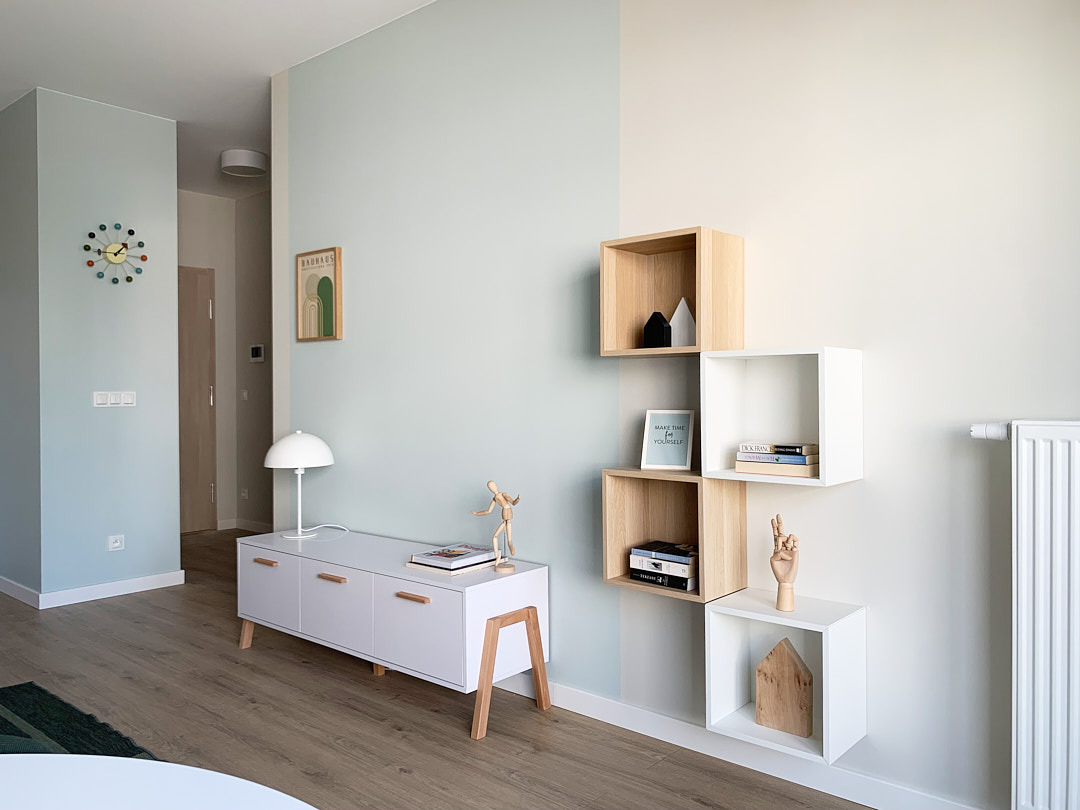 Jasne wnętrze: biała komoda z drewnianymi nogami, lampka, dekoracje, modułowe półki na ścianie, zegar w stylu retro, plakat Bauhaus. Minimalistyczny design.