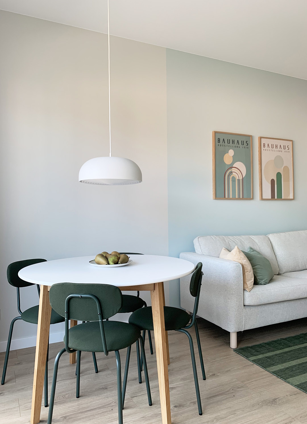 Jasne wnętrze: okrągły stół z gruszkami, zielone krzesła, sofa z poduszkami, lampa wisząca i plakaty Bauhaus na ścianie. Minimalistyczny design.