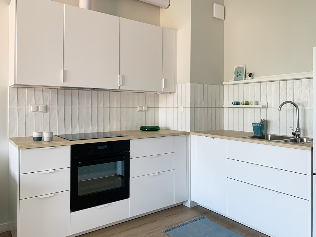 Jasna kuchnia w stylu skandynawskim: białe szafki z drewnianym blatem, pionowe płytki na ścianie, minimalistyczny wystrój i subtelny dywanik.