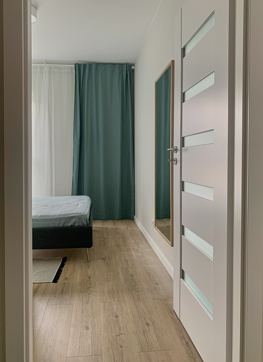 Nowoczesna sypialnia z łóżkiem, turkusowymi zasłonami i lustrem w złotej ramie, widziana zza uchylonych, białych drzwi z przeszkleniami.