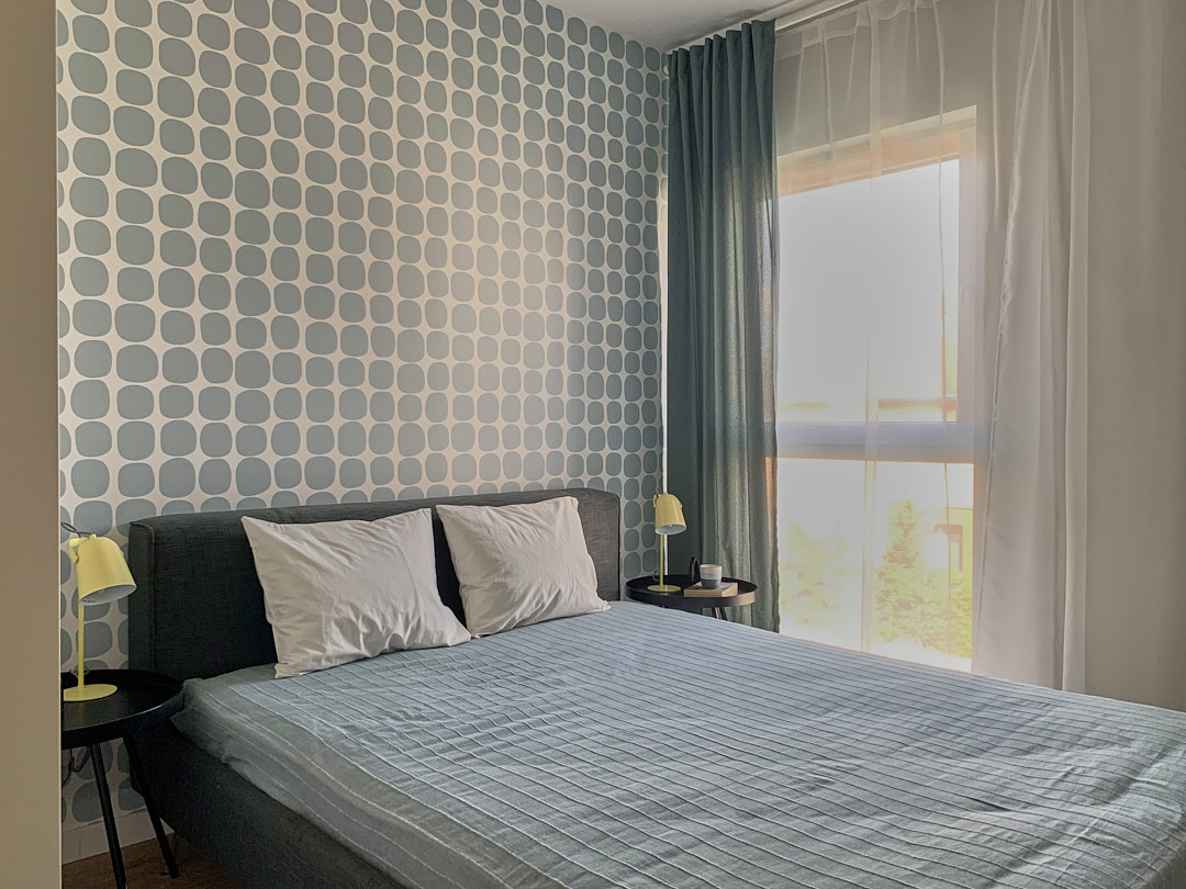 Sypialnia z łóżkiem w szarej ramie, niebieską pościelą i tapetą w geometryczne wzory. Okno z zasłonami w odcieniach bieli i szarości. Żółte lampki nocne na stolikach.