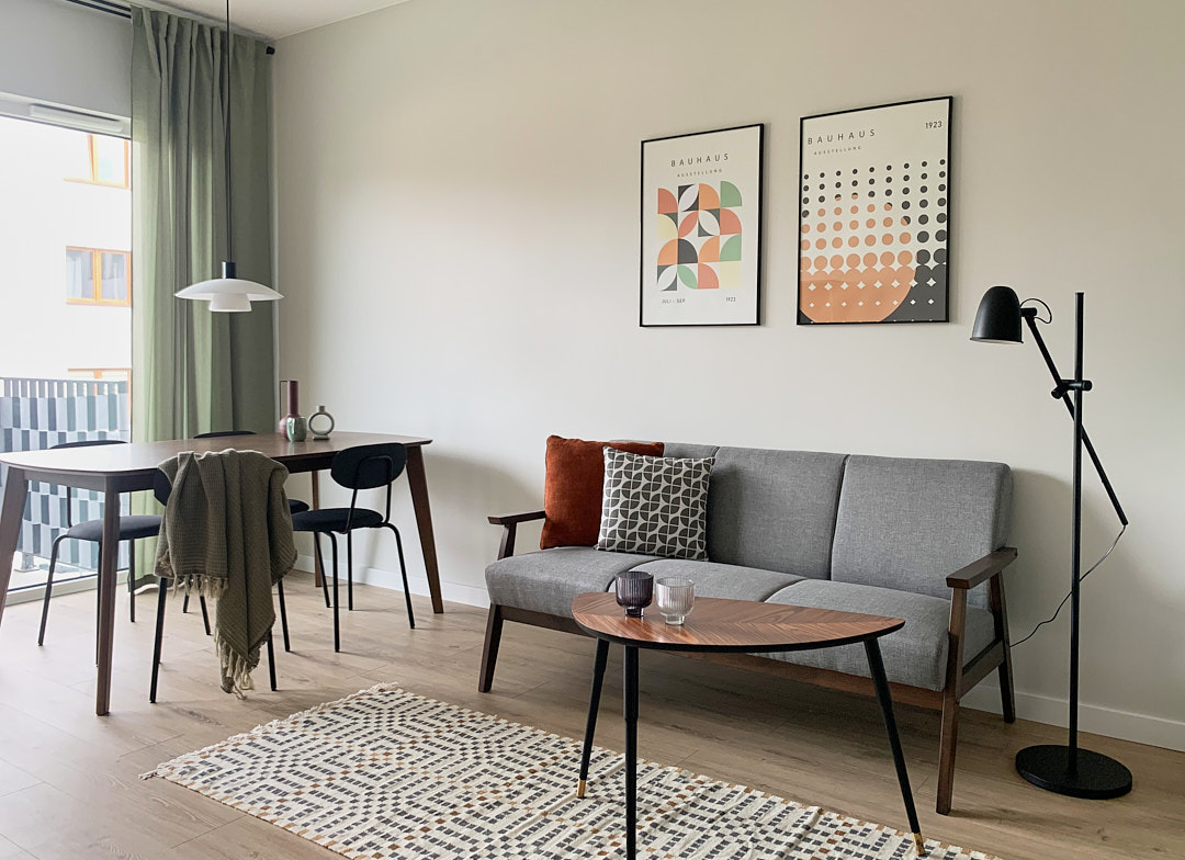 Jasne wnętrze w stylu Bauhaus: szara sofa, drewniany stół, geometryczny dywan i plakaty na ścianie. Minimalistyczna lampa podłogowa i stonowana kolorystyka.