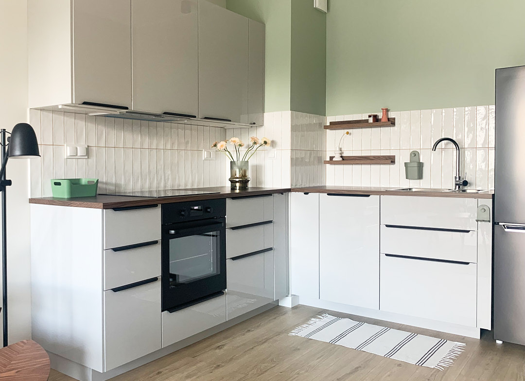 Jasna kuchnia w stylu skandynawskim: białe szafki z czarnymi uchwytami, drewniany blat, płytki na ścianie i zielony akcent kolorystyczny. Minimalistyczny design.