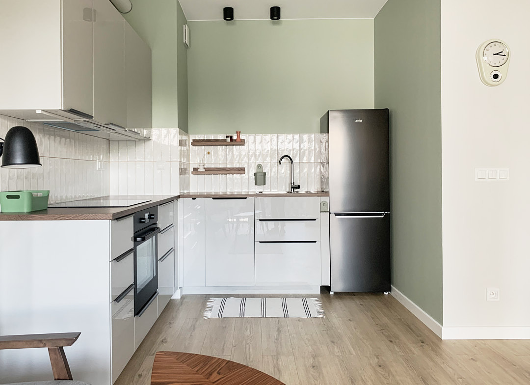 Nowoczesna kuchnia w odcieniach bieli i zieleni z drewnianymi akcentami. Widoczne błyszczące fronty szafek, czarny sprzęt AGD i dekoracyjne półki nad blatem.