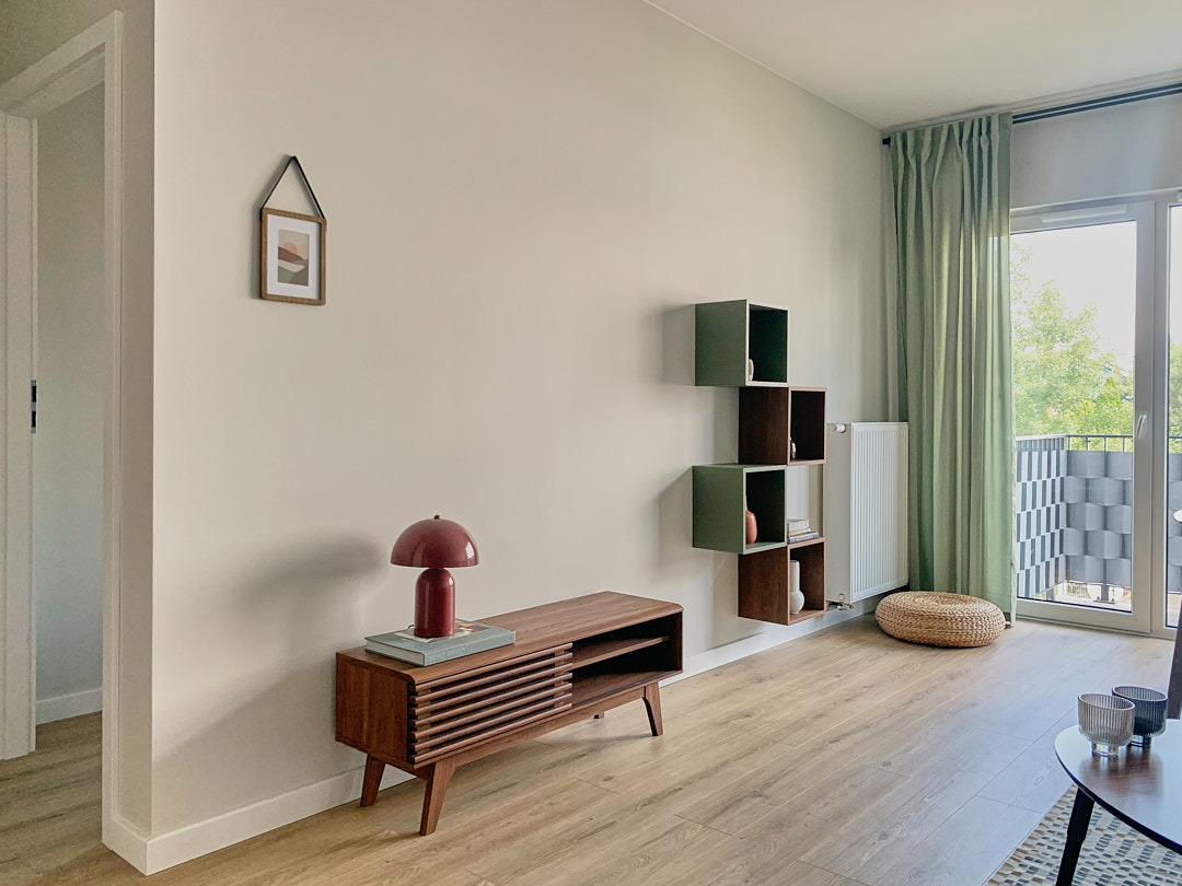 Jasne wnętrze z drewnianą komodą, lampką w stylu retro i modułowymi półkami na ścianie. Uzupełnia je grafika w ramce i zielone zasłony.