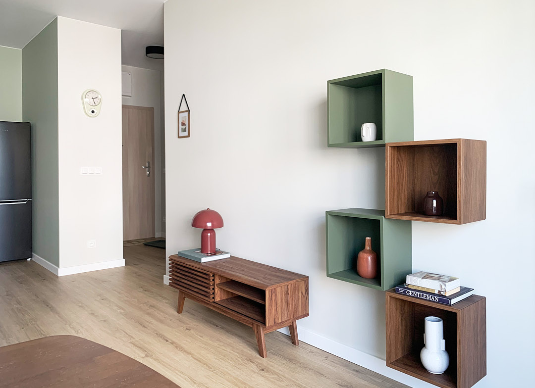 Nowoczesny salon z drewnianą komodą, czerwoną lampką i asymetrycznymi półkami w odcieniach zieleni i brązu. Minimalistyczny wystrój, jasne drewno na podłodze.