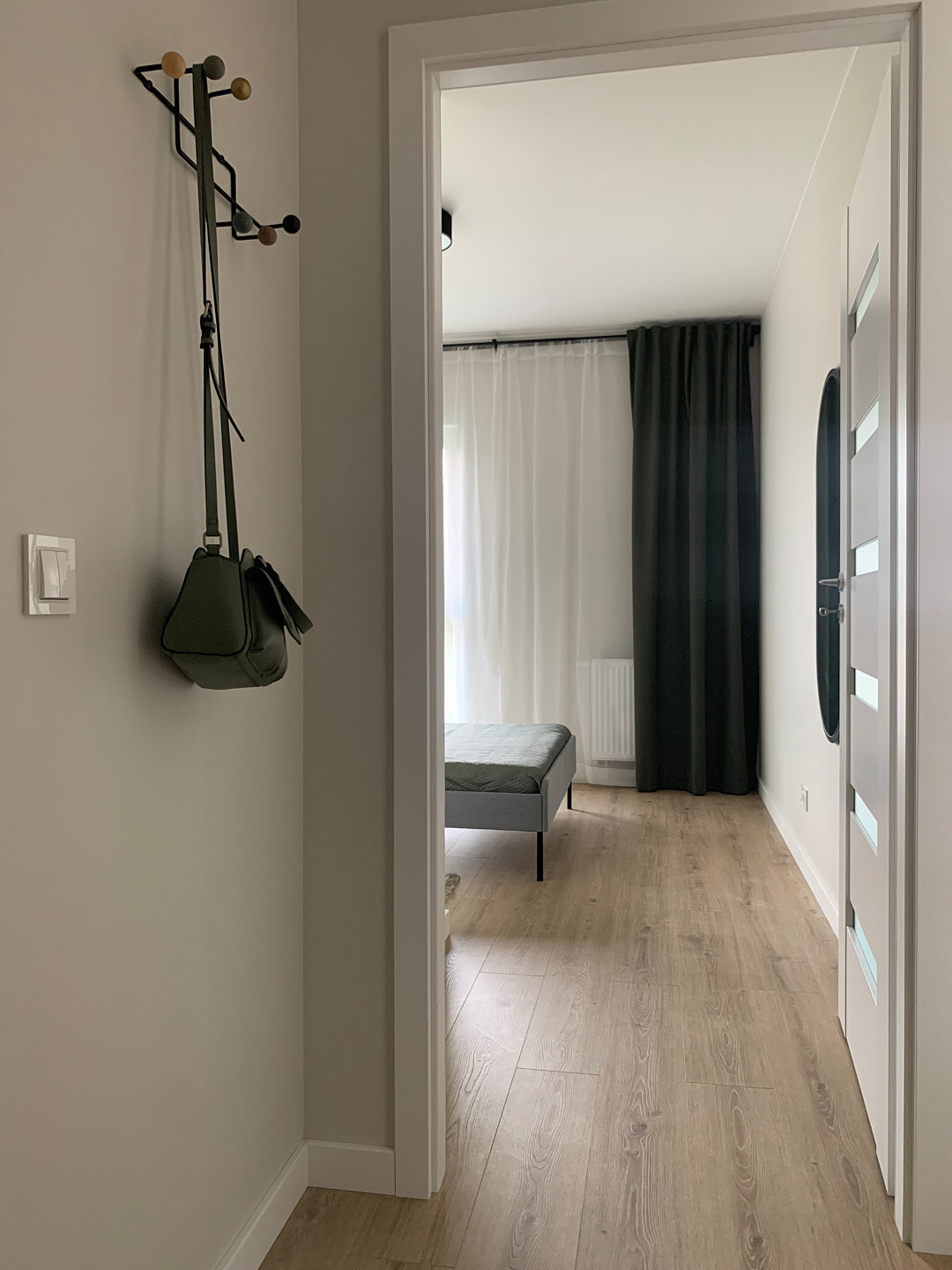 Nowoczesny przedpokój z drewnianą podłogą, wieszakiem na ścianie i widokiem na pokój z szarą ławką i zasłonami. Minimalistyczny design, jasne wnętrze.