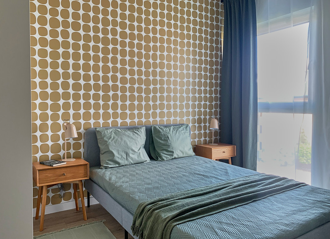 Sypialnia z łóżkiem w odcieniach szarości i zieleni, wzorzysta tapeta w stylu retro, drewniane szafki nocne z lampkami i zasłony wpadające do pokoju przez okno.