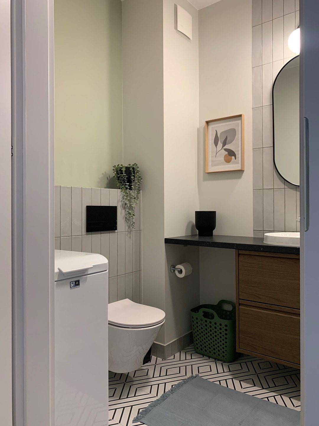 Nowoczesna łazienka w odcieniach szarości i zieleni z geometryczną podłogą, wiszącą toaletą i drewnianą szafką pod umywalką. Uwagę przykuwa roślina w czarnej doniczce.