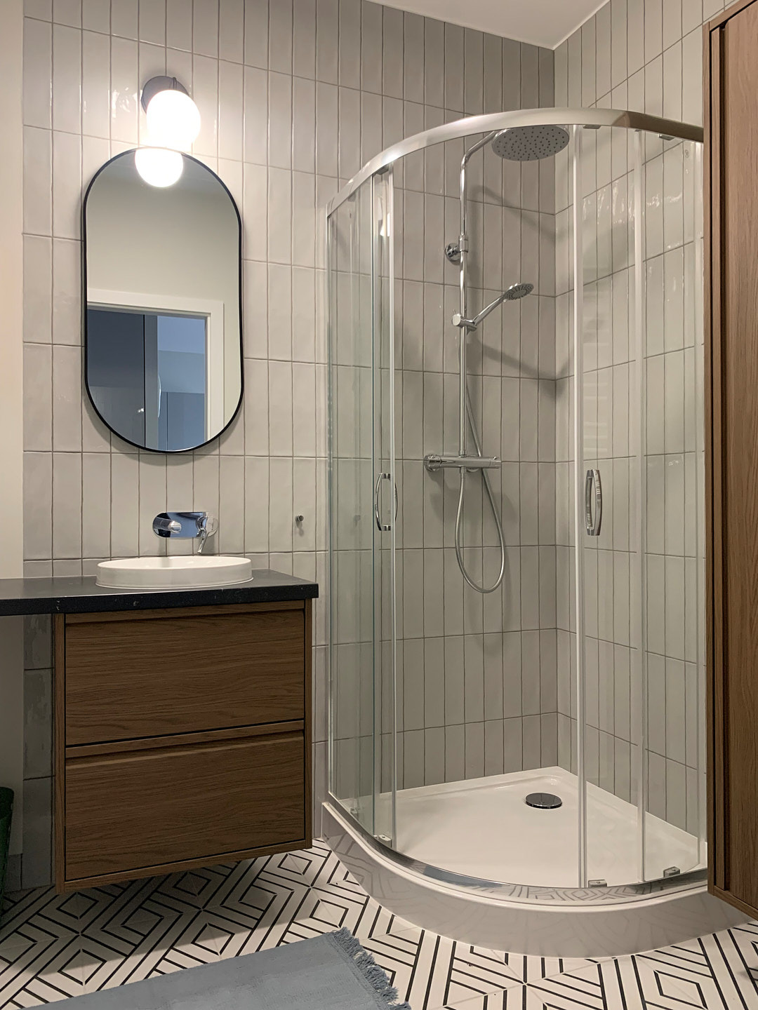 Nowoczesna łazienka z kabiną prysznicową narożną, umywalką na szafce z ciemnym blatem i owalnym lustrem podświetlanym. Geometryczny wzór na podłodze.