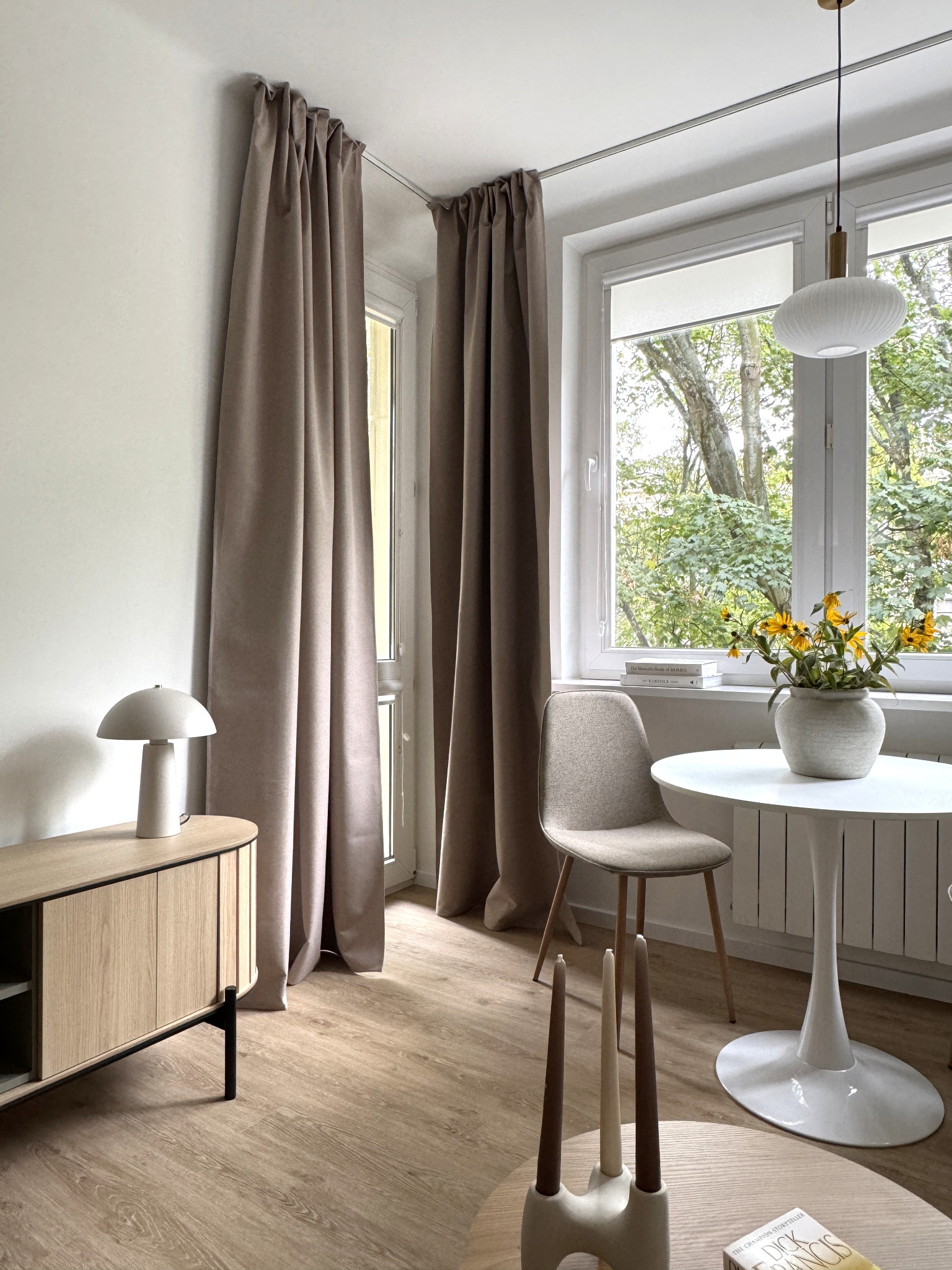 Jasne wnętrze z beżowymi zasłonami, drewnianą komodą i okrągłym stołem z kwiatami. Minimalistyczny design, naturalne światło, widok na zieleń za oknem.