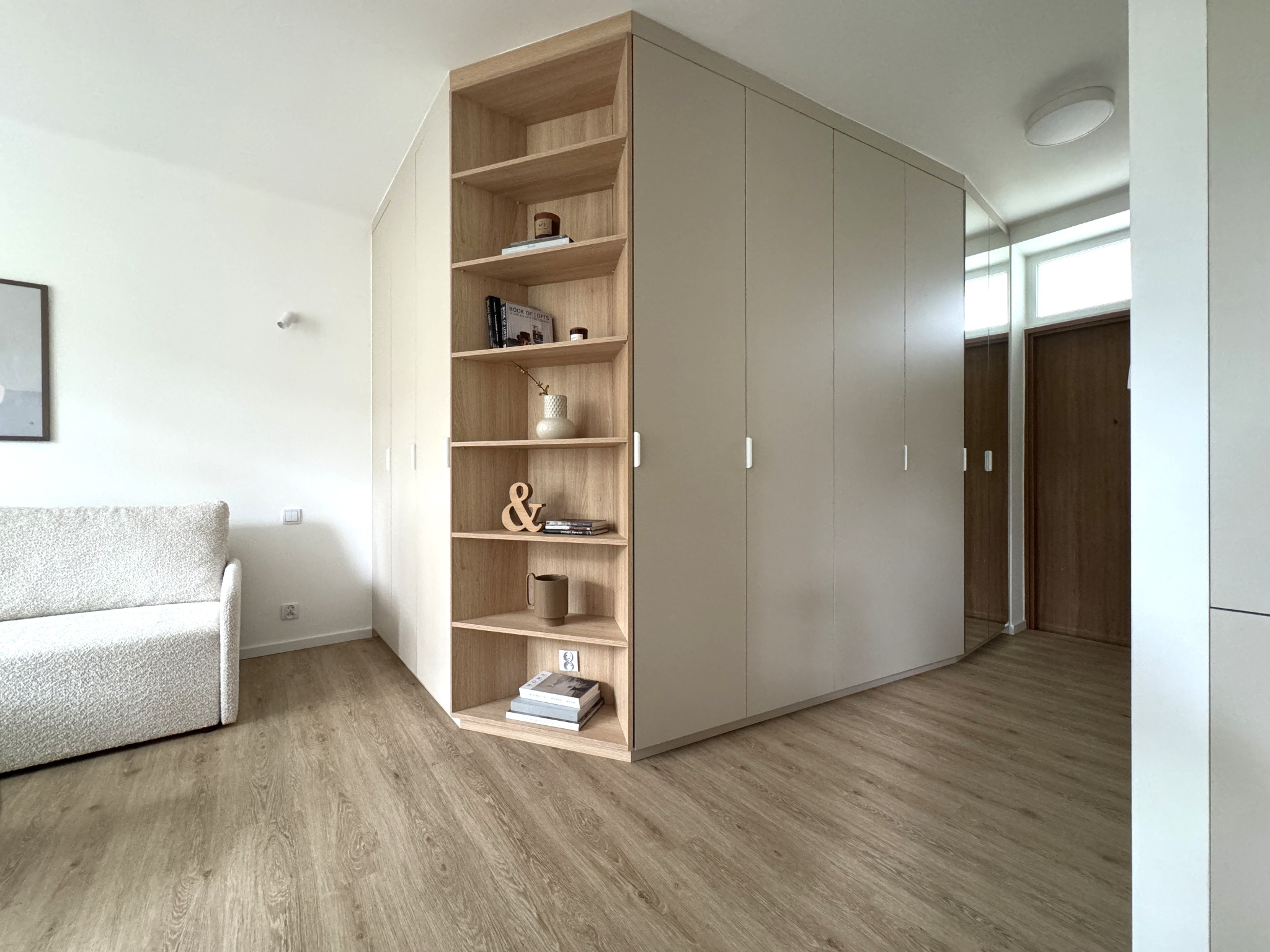 Biało-beżowa zabudowa meblowa z półkami w rogu pokoju, na których stoją książki i dekoracje. Jasna podłoga i sofa w tle. Minimalistyczny styl.