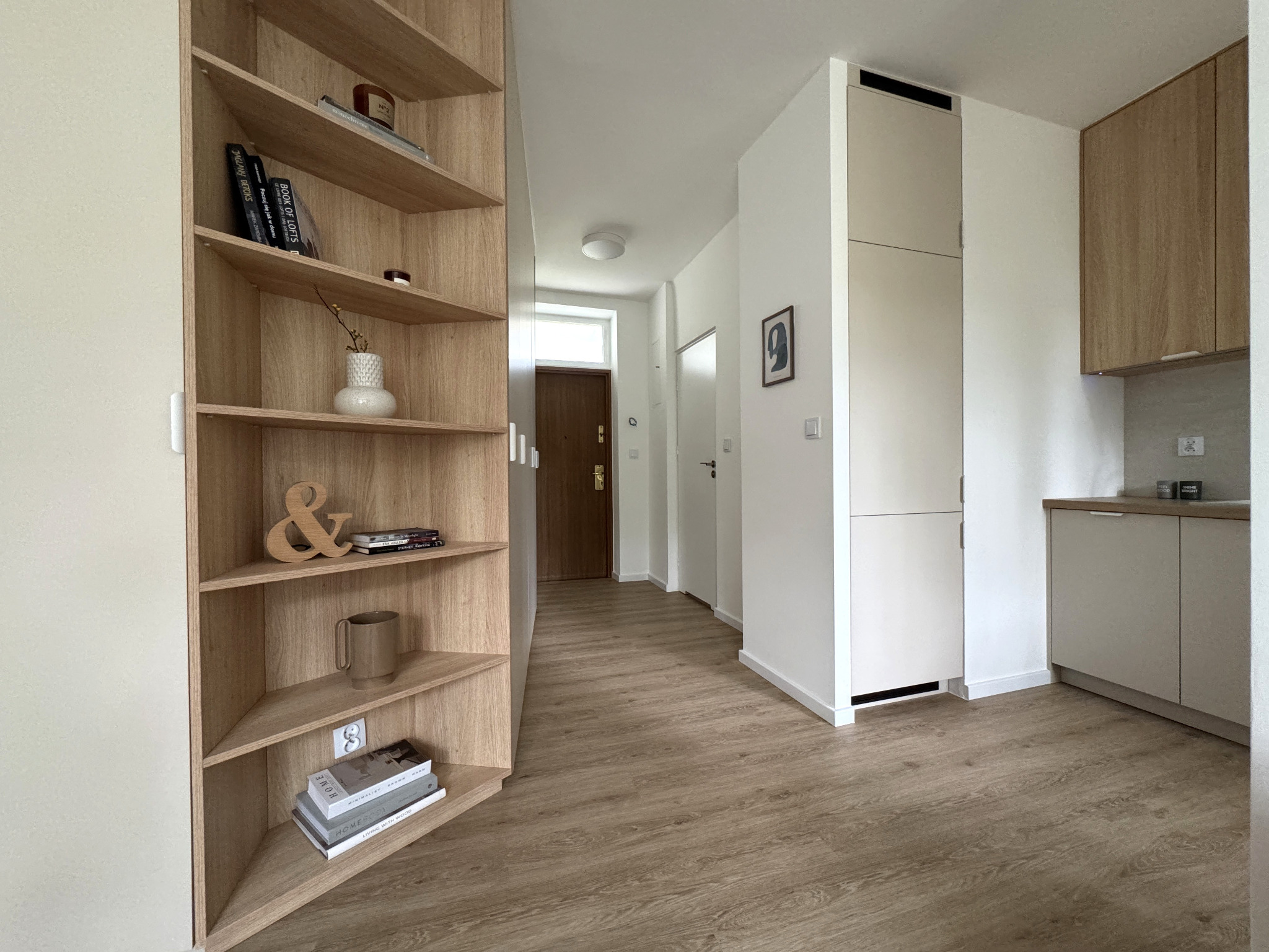Jasne wnętrze mieszkania z drewnianą podłogą i regałem narożnym. W tle widoczny korytarz i fragment kuchni. Minimalistyczny design z elementami dekoracyjnymi.