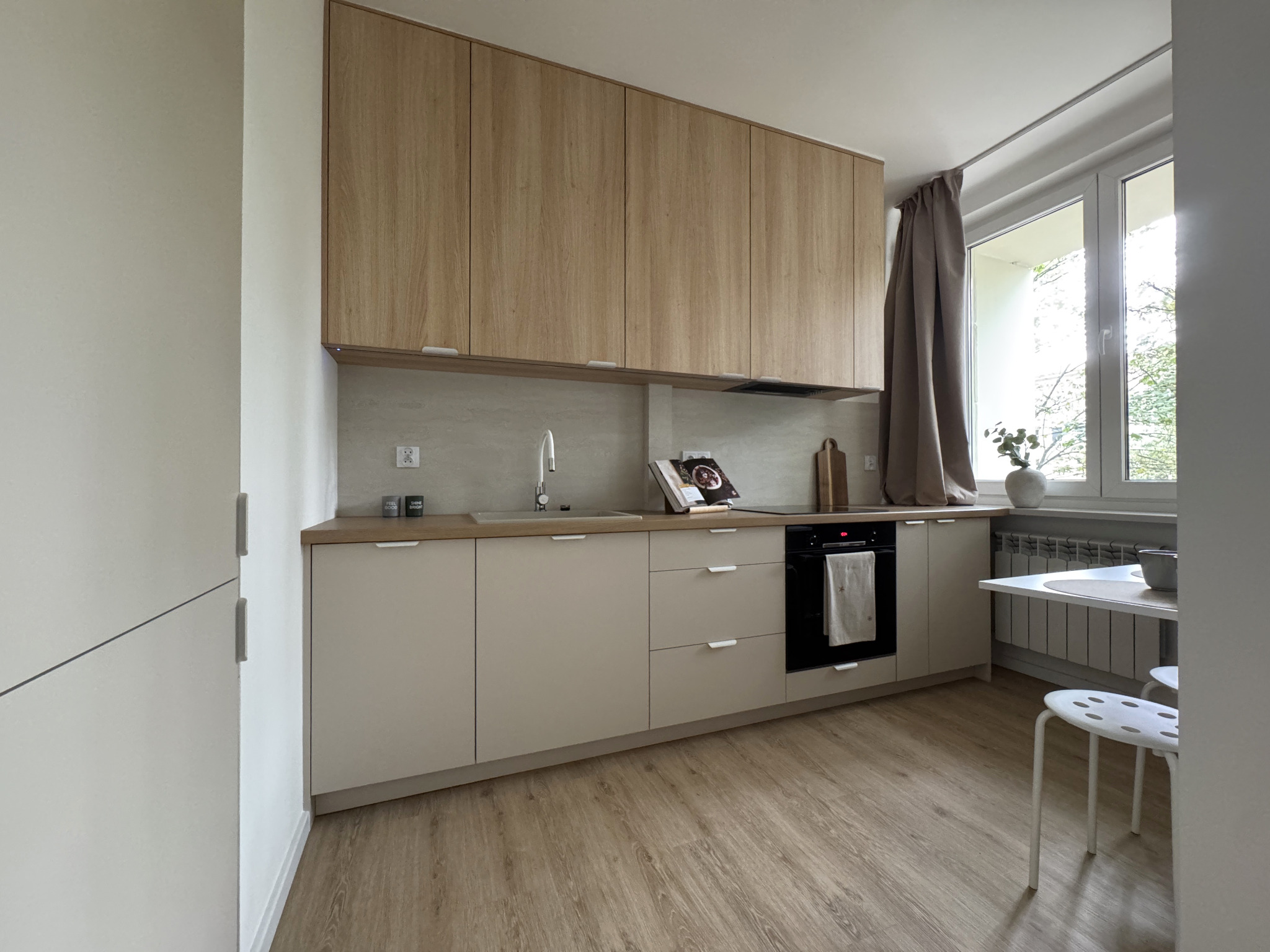 Jasna kuchnia z drewnianymi szafkami górnymi i beżowymi dolnymi. Na blacie książka kucharska i deska do krojenia. Biały stół z krzesłami przy oknie z zasłoną.