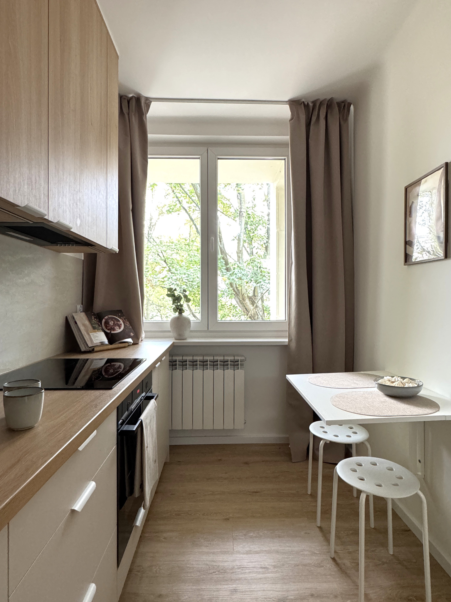 Jasna, wąska kuchnia z drewnianymi akcentami: zabudowa, blat, podłoga. Biały stół z krzesłami, okno z widokiem na zieleń. Minimalistyczny wystrój.