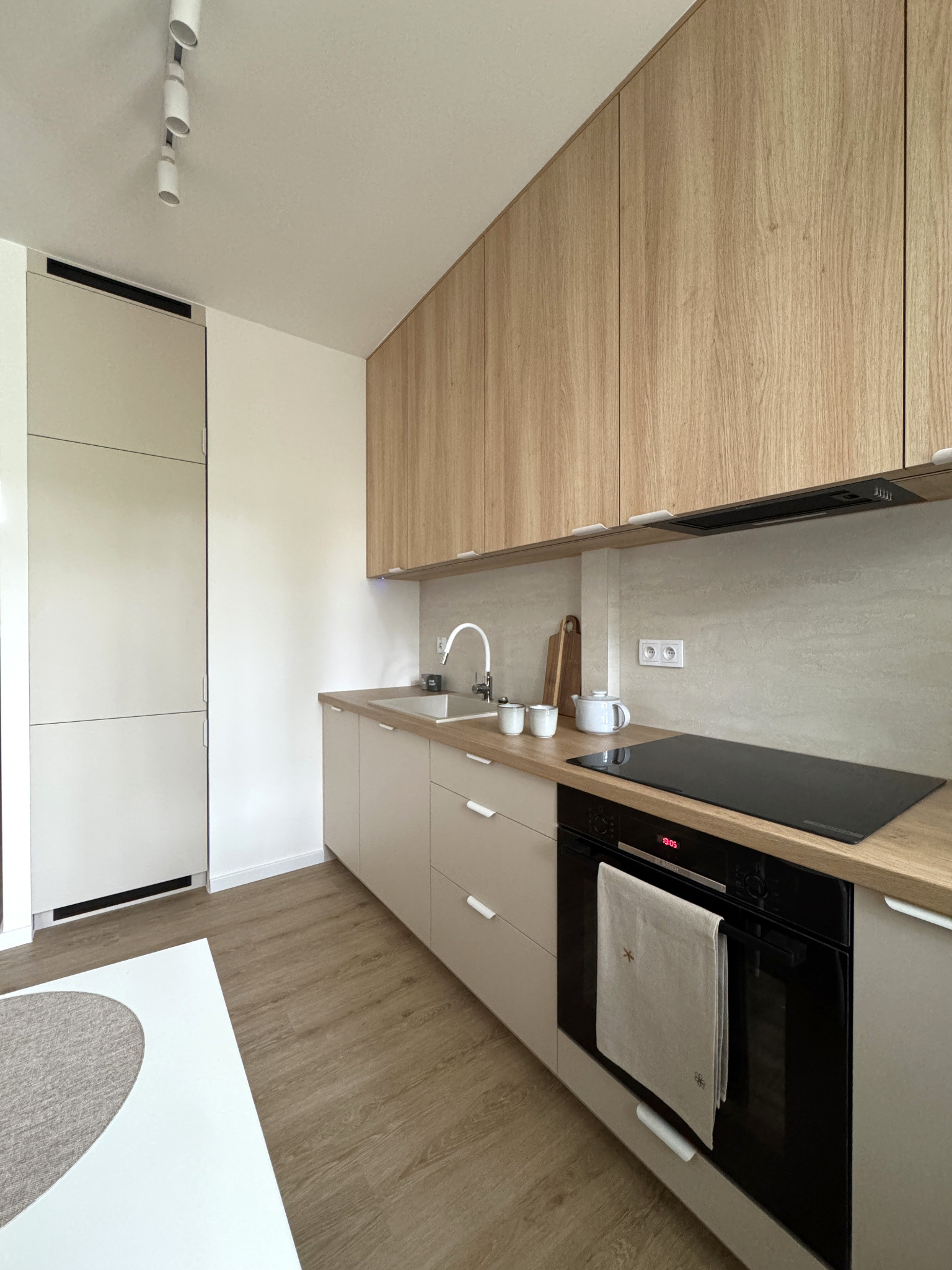 Jasna kuchnia w stylu skandynawskim: drewniane szafki górne, beżowe dolne, czarna płyta indukcyjna i piekarnik. Minimalistyczne oświetlenie sufitowe.