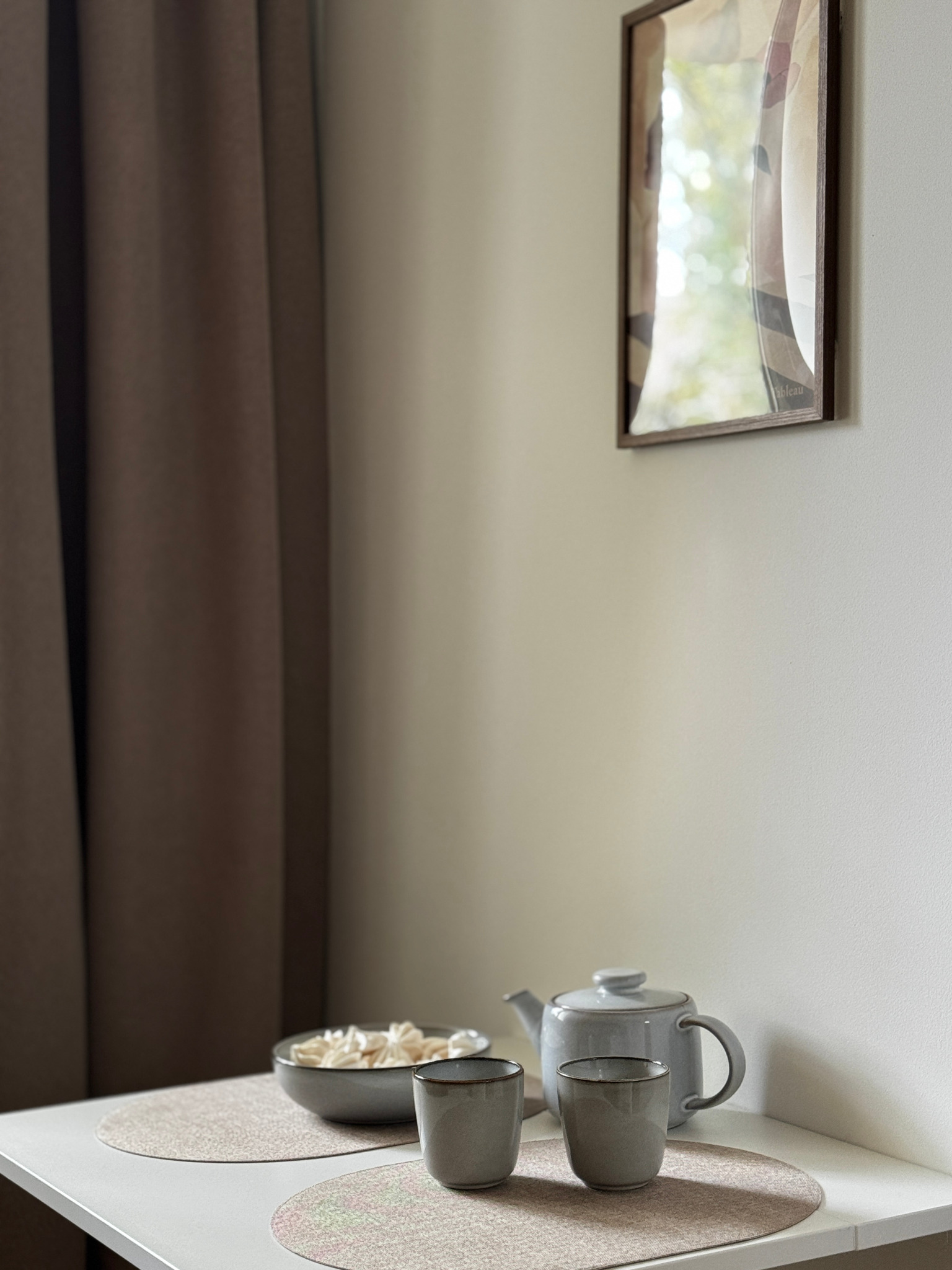 Minimalistyczna aranżacja stołu z szarą zastawą, misą z bezami i obrazem w ramie na ścianie. Stonowana kolorystyka, naturalne światło, przytulny kącik w mieszkaniu.