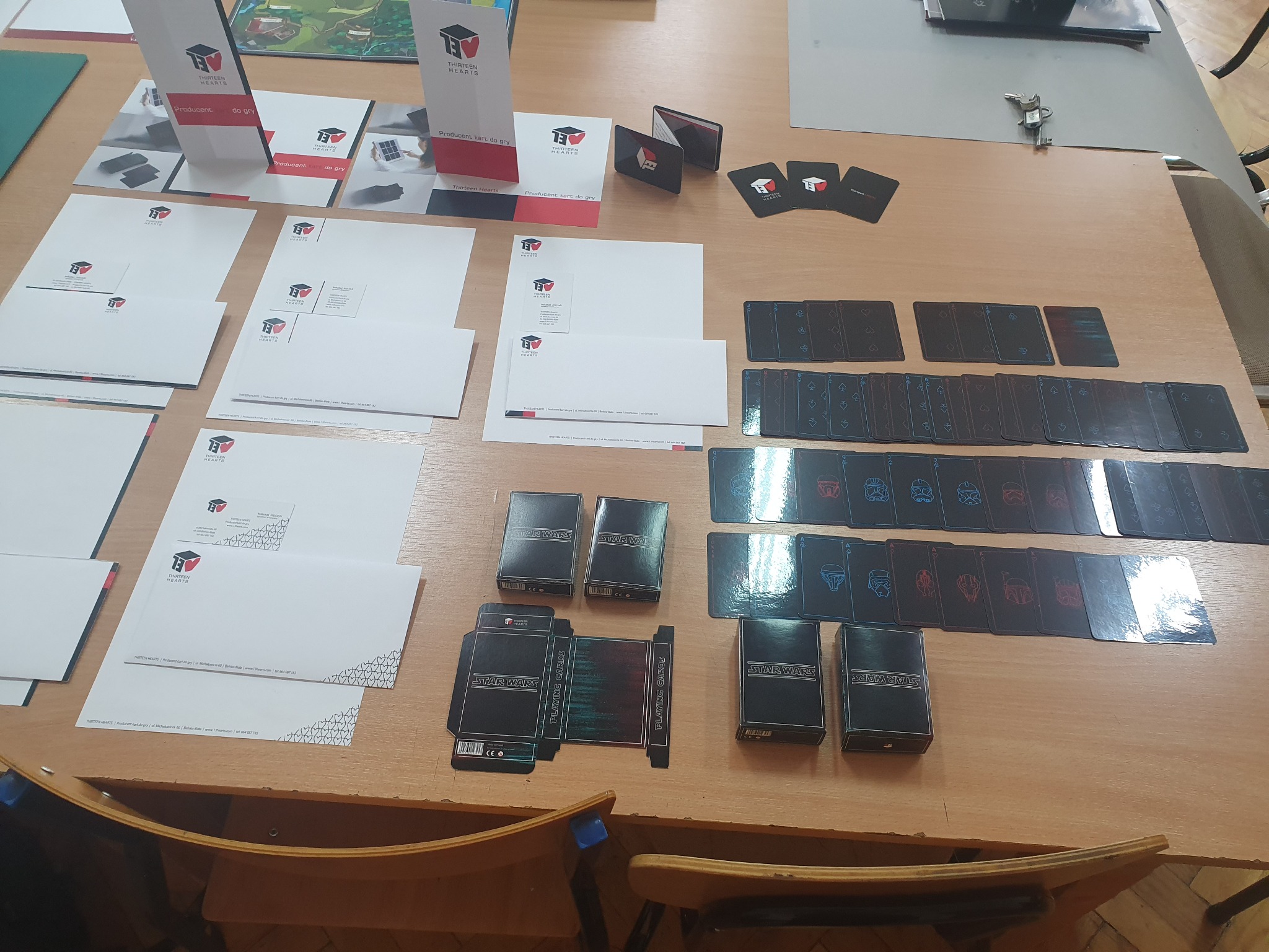 Stół z rozłożonymi elementami identyfikacji wizualnej firmy Thirteen Hearts, w tym wizytówki, papier firmowy, ulotki, oraz talie kart do gry Star Wars w czarnych pudełkach i rozłożone karty...