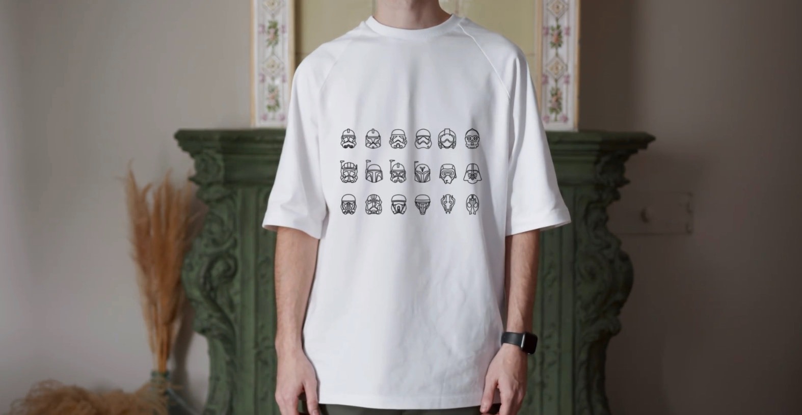 Mężczyzna w białym T-shircie z czarnymi grafikami hełmów postaci z Gwiezdnych Wojen, stojący na tle kominka z dekoracjami.