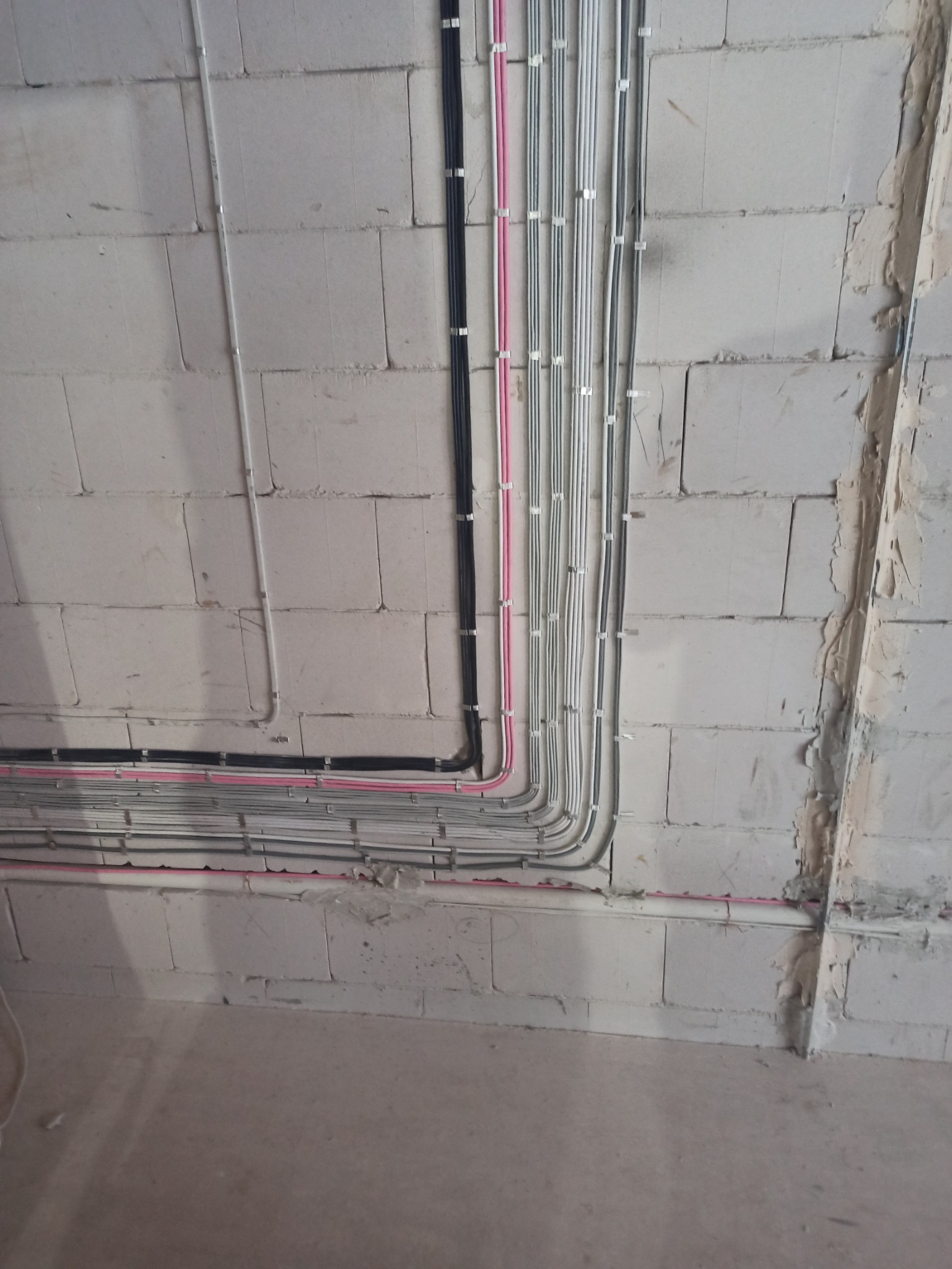 Ściana z betonu komórkowego z poprowadzonymi przewodami elektrycznymi w peszlach i listwach, widoczne przewody czarne, białe, szare i różowe.