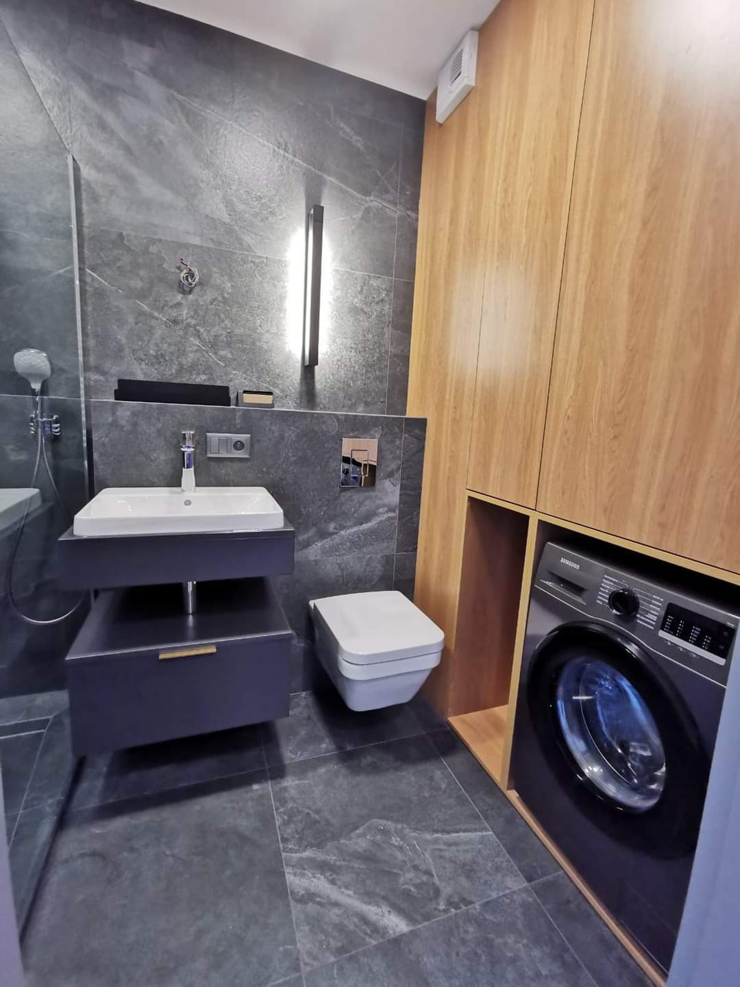 Nowoczesna łazienka z szarymi płytkami imitującymi kamień, podwieszana szafka z umywalką, toaleta i pralka zabudowana w drewnianej szafie.