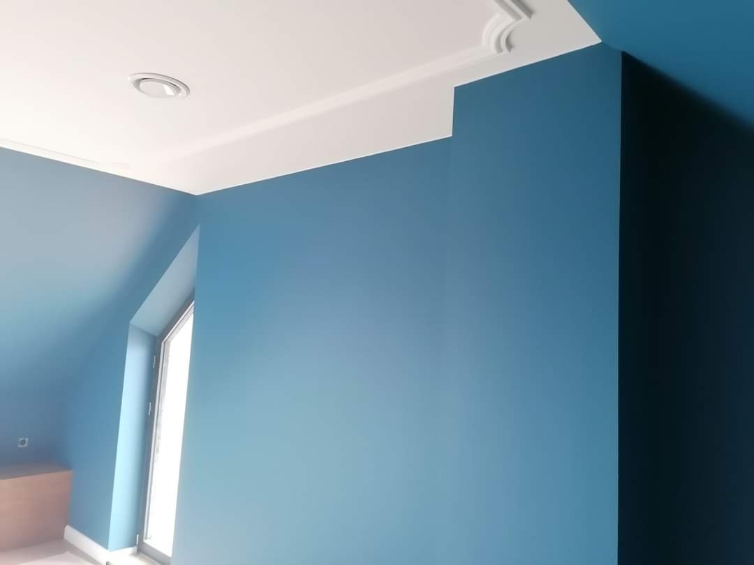 Pomieszczenie po malowaniu na odcienie niebieskiego, z białym sufitem i listwami ozdobnymi, widoczne okno i fragment mebla.