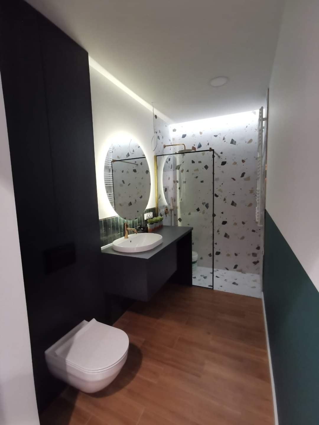 Nowoczesna łazienka z drewnopodobną podłogą, czarną ścianą z podwieszaną toaletą i umywalką na szafce, okrągłe lustro z podświetleniem LED oraz kabina prysznicowa z mozaiką w odcieniach szarości...