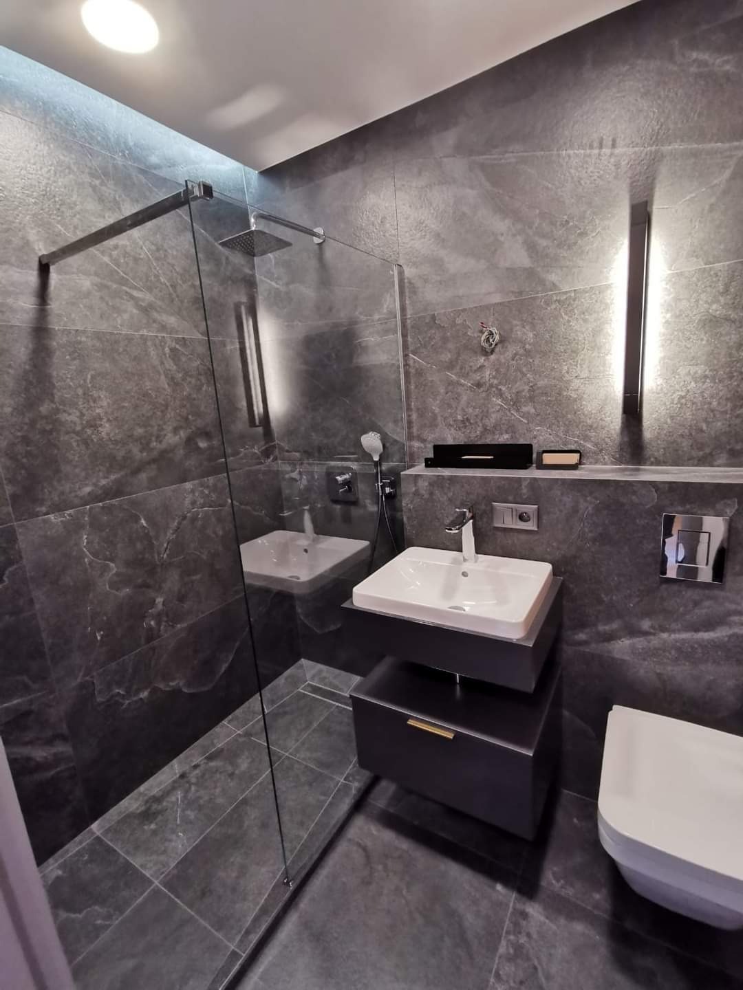 Nowoczesna łazienka z ciemnymi, marmurkowymi płytkami, kabiną prysznicową ze szklaną ścianką, wiszącą szafką z umywalką i podświetleniem LED na ścianie.