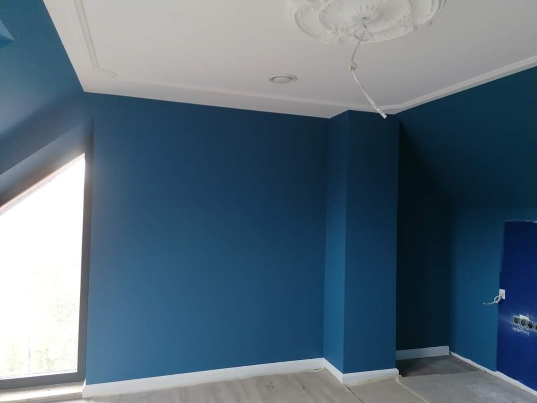 Pomieszczenie po remoncie z pomalowanymi na niebiesko ścianami, białym sufitem z dekoracyjną rozetą i wnęką okienną.