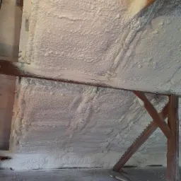 Poddasze z drewnianą konstrukcją pokryte grubą warstwą białej piany izolacyjnej, widoczne niedociągnięcia i nierówności w aplikacji, surowy betonowy podkład.