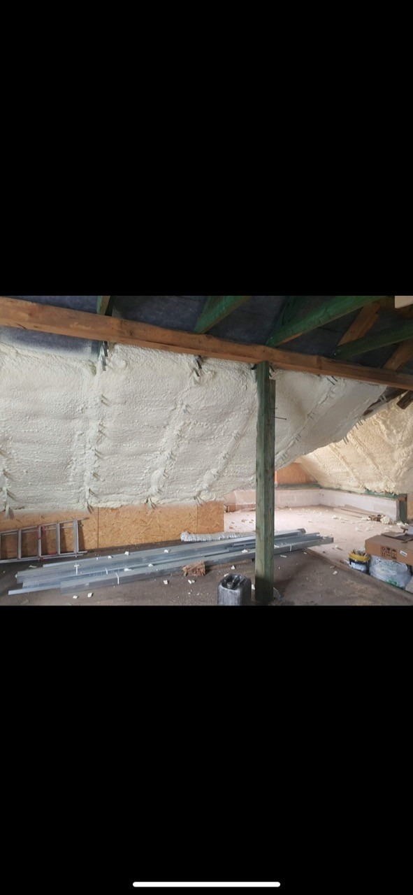 Poddasze z widoczną warstwą białej pianki izolacyjnej na skośnych ścianach i suficie, drewniane elementy konstrukcyjne dachu, rozłożone narzędzia i materiały budowlane na podłodze.